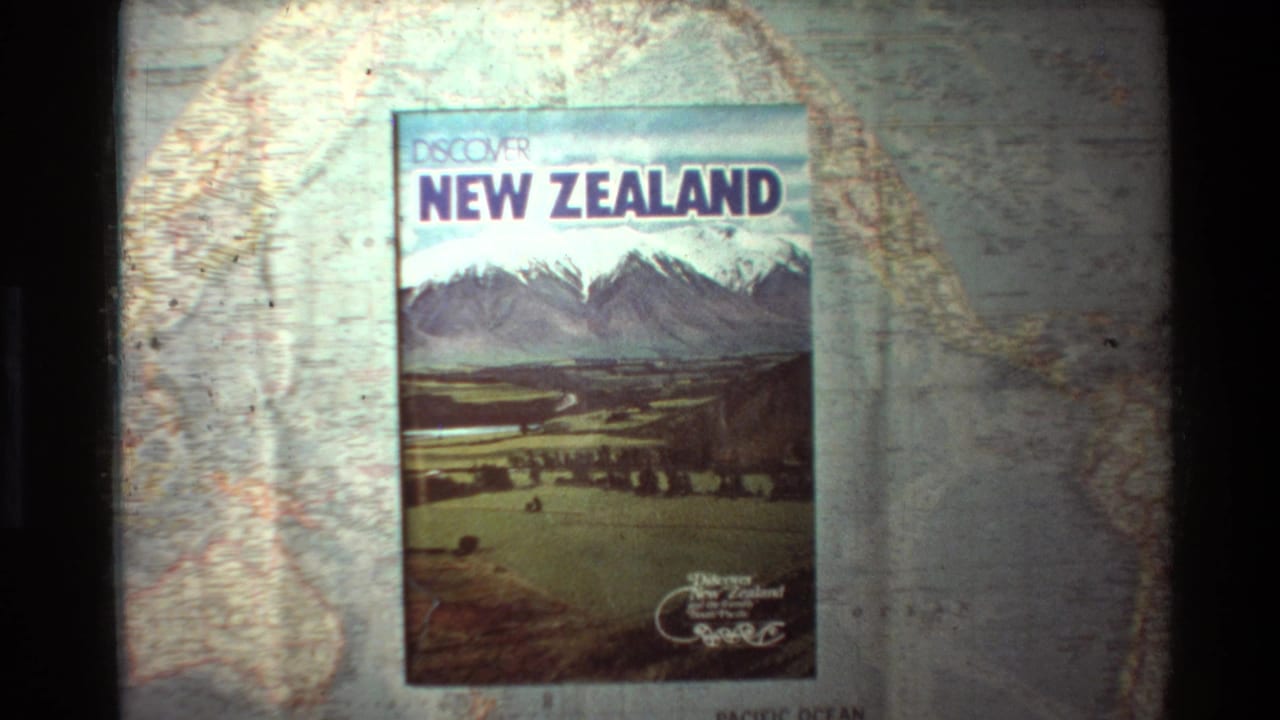 1981 8mm frame showing a Discover New Zealand travel book on a printed map, with mountain cover art and visible film grain.