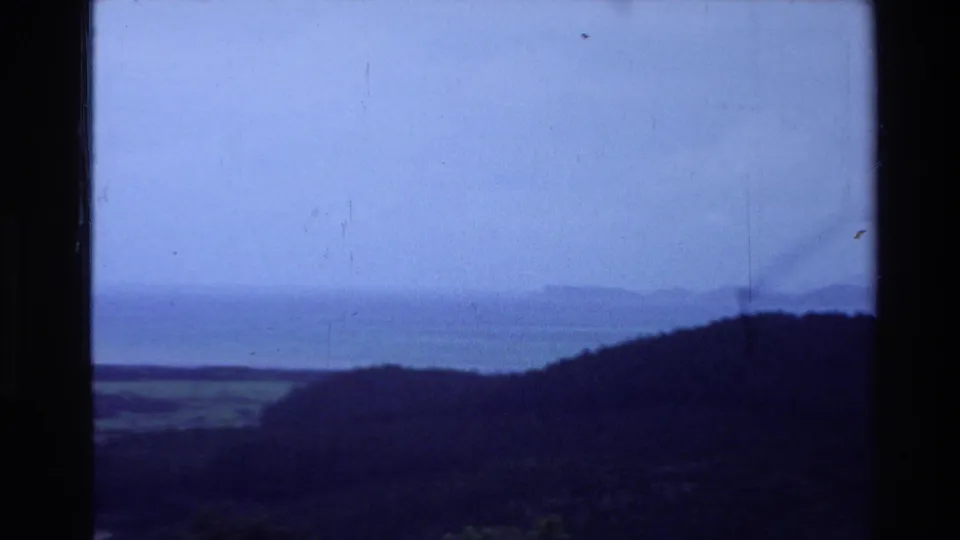 A cool blue New Zealand coastal view at dusk, with dark hills in the foreground and distant water on the horizon.
