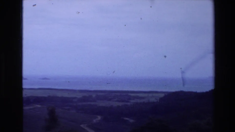 A wide New Zealand landscape at dusk shows hills, open fields, distant water, and a pale winding river or road.