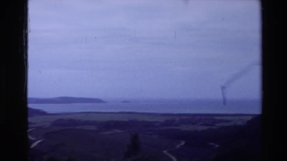 1981 New Zealand 8mm frame showing distant water, low hills, cloudy sky, and a purple-blue film cast.