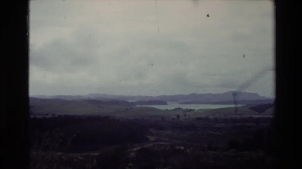 1981 New Zealand home-movie frame showing a broad sea view, foreground hillside, distant islands, and overcast sky.