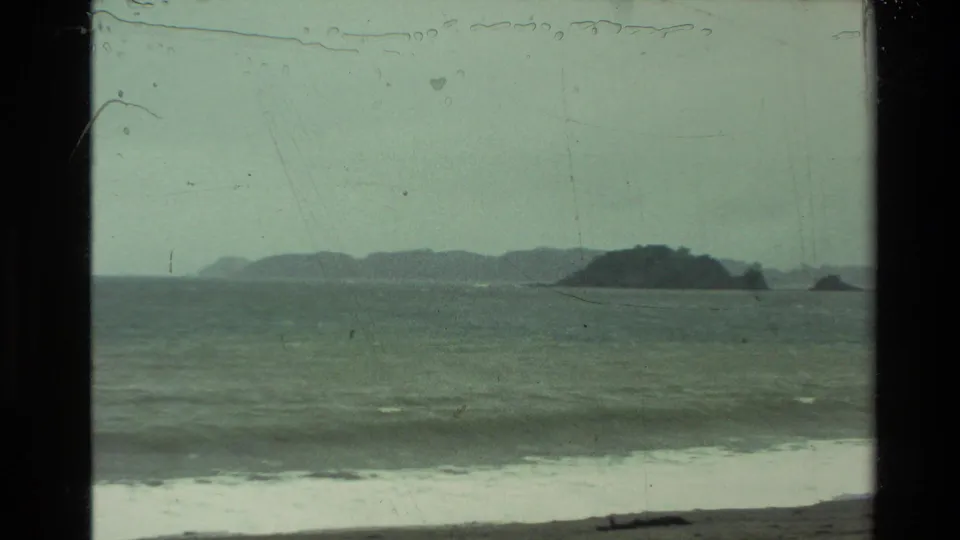 1981 New Zealand 8mm frame showing gray surf, a dark shoreline, and several distant rocky outcrops under an overcast sky.