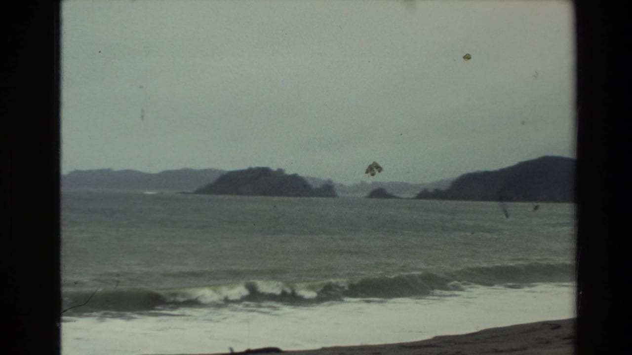 1981 New Zealand coastal frame with a breaking wave, muted sea, offshore rocks, and hazy hills seen through aged 8mm film texture.