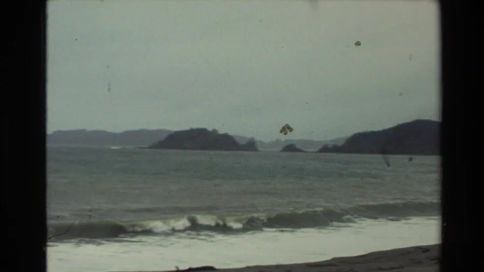 1981 New Zealand coastal frame with a breaking wave, muted sea, offshore rocks, and hazy hills seen through aged 8mm film texture.