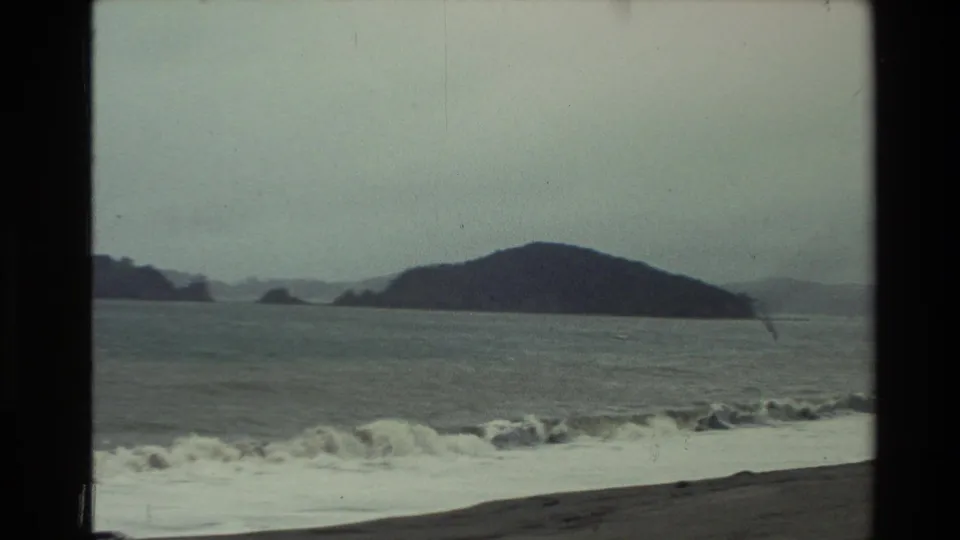 1981 New Zealand 8mm frame showing foamy rough surf, a sloping dark beach, and a large offshore rock form in the distance.
