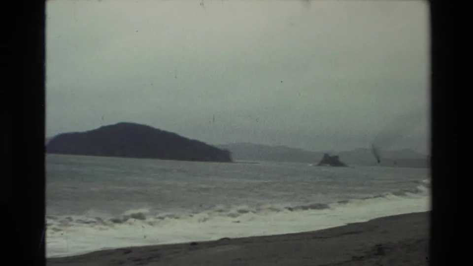 1981 New Zealand 8mm frame with rough gray water, a dark foreground beach, distant hills, and small rocky outcrops under cloud cover.