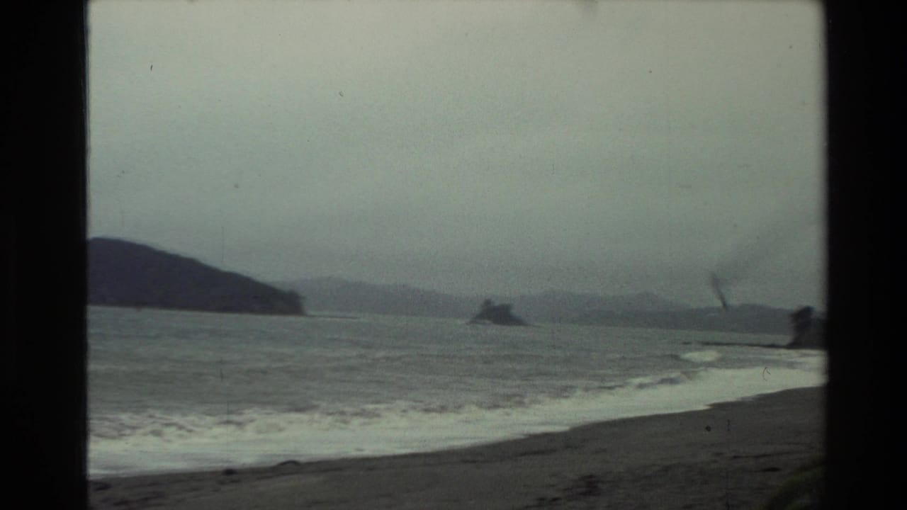 1981 New Zealand home-movie view of an overcast coast, low surf, distant hills, and a small offshore landform framed by dark 8mm borders.
