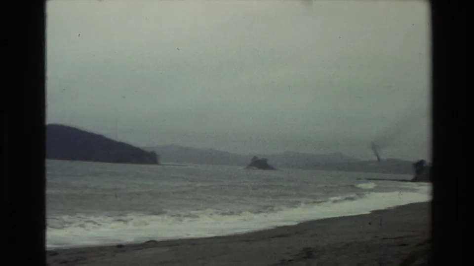 1981 New Zealand home-movie view of an overcast coast, low surf, distant hills, and a small offshore landform framed by dark 8mm borders.
