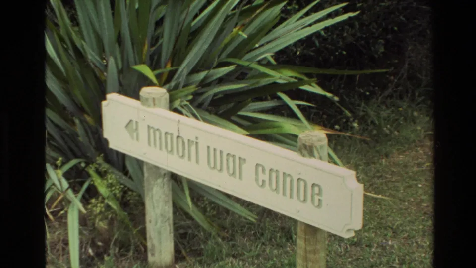 Wooden directional sign reading Maori War Canoe beside dense green plants, filmed in New Zealand on 8mm home-movie stock.