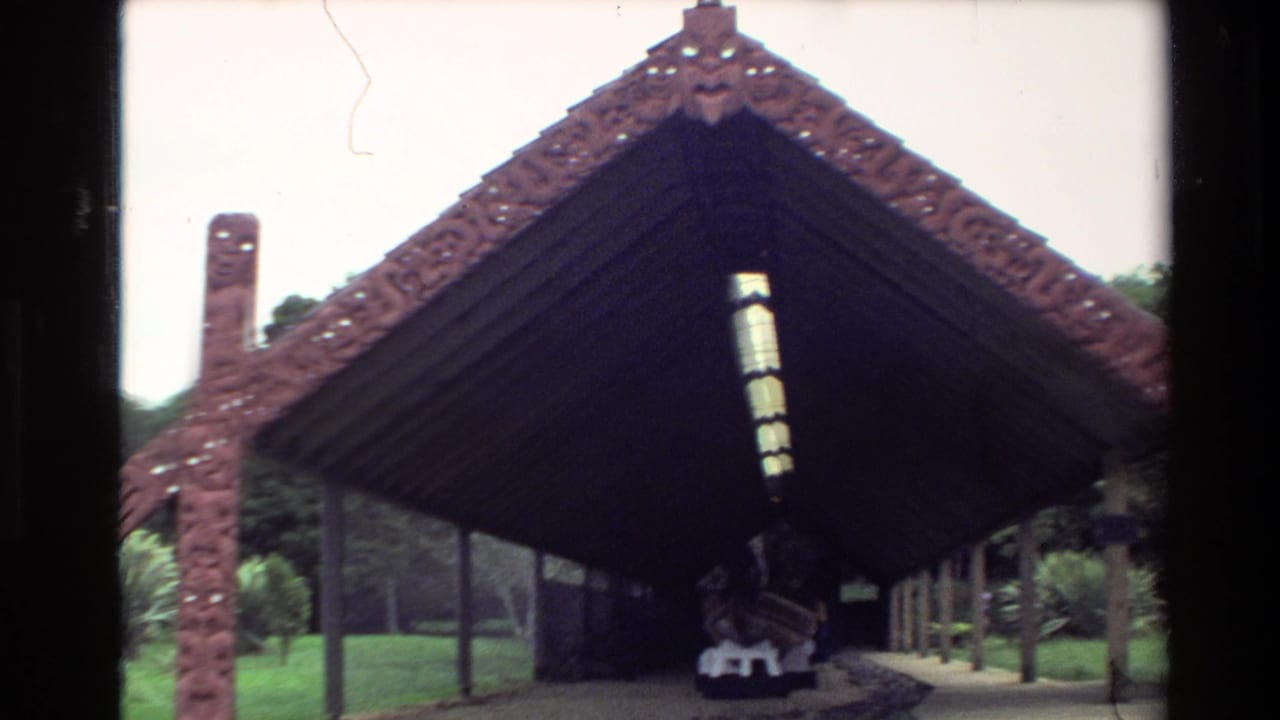 1981 New Zealand home-movie frame showing a carved wooden shelter with a traditional waka displayed under the dark roof.