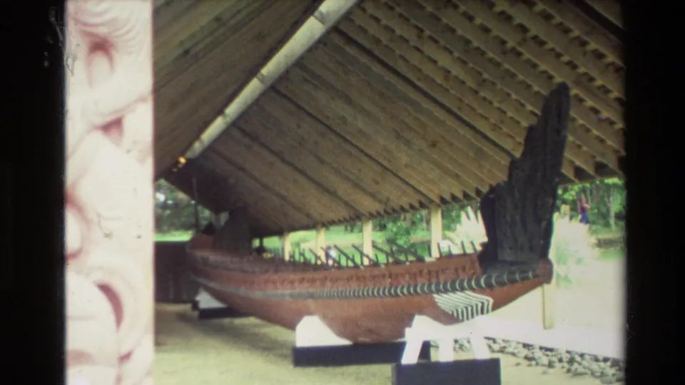Closer 1981 New Zealand home-movie view of a waka resting on supports under a carved wooden meeting-house shelter.