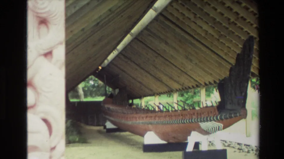 1981 New Zealand frame showing the waka from the side under roof beams, with carved architectural detail at the edge.