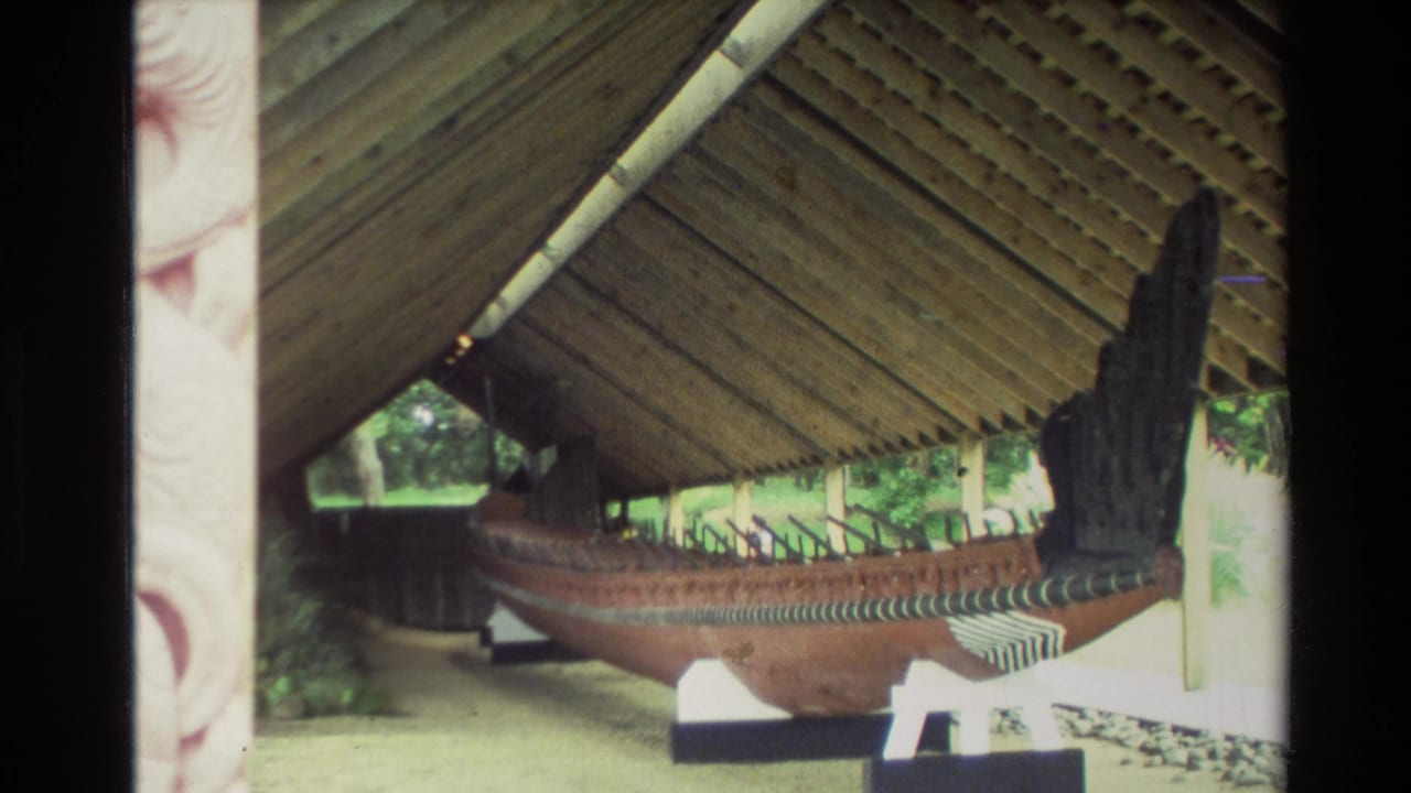 1981 New Zealand home-movie frame showing a large wooden waka under a timber shelter, viewed from the side.