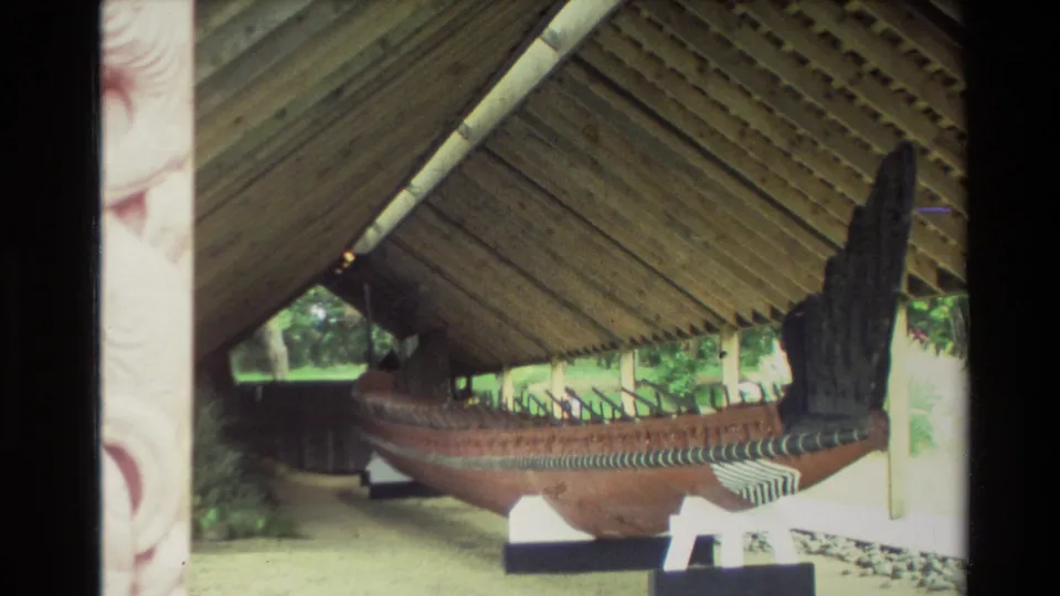 1981 New Zealand home-movie frame showing a large wooden waka under a timber shelter, viewed from the side.