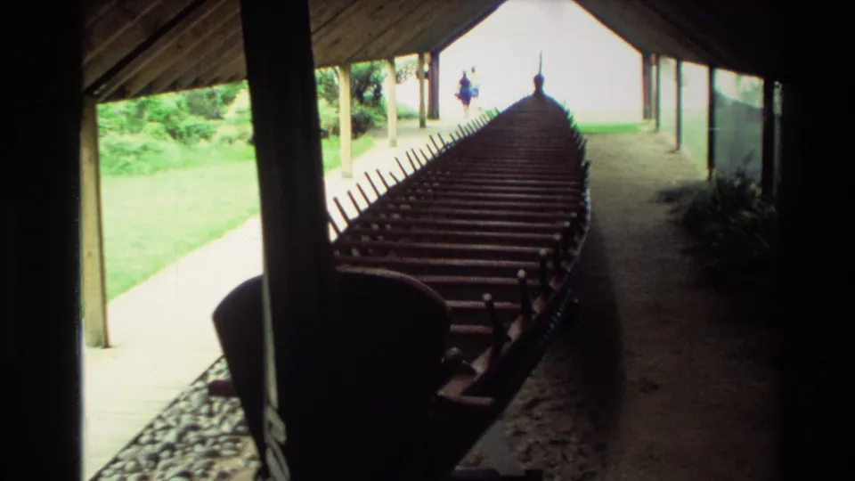 1981 New Zealand frame looking along a long wooden waka with repeated seats beneath a covered display area.