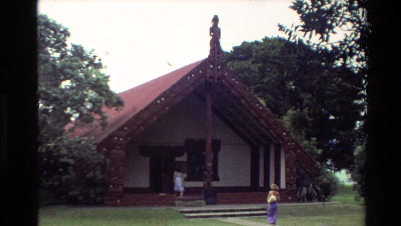 1981 New Zealand home-movie frame showing a Māori meeting house exterior with carved gable details, red roof and visitors nearby.