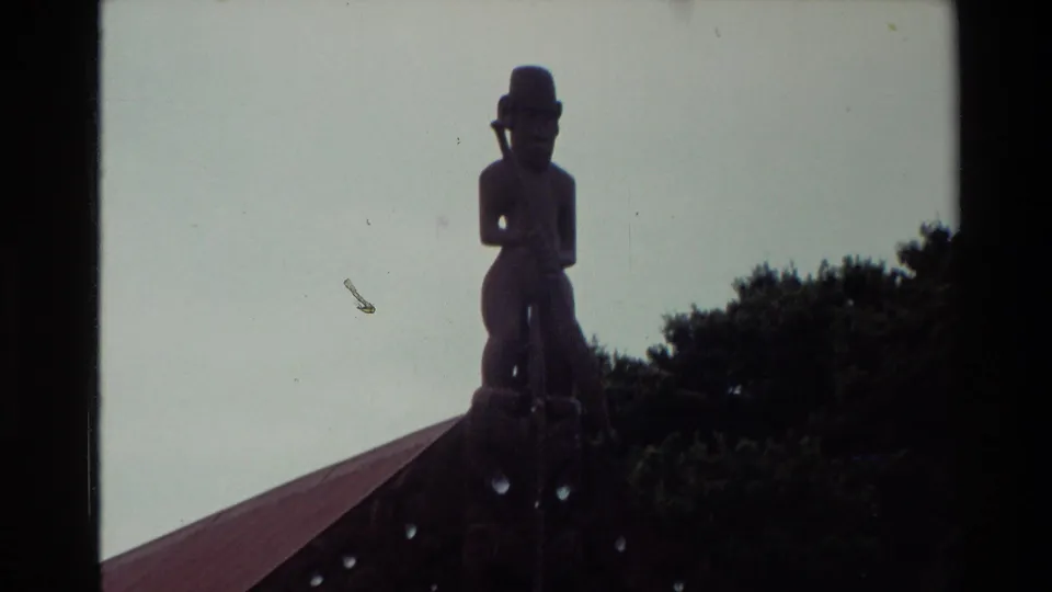 Tight 1981 New Zealand home-movie view of a carved figure on the meeting-house roofline, framed against pale sky.