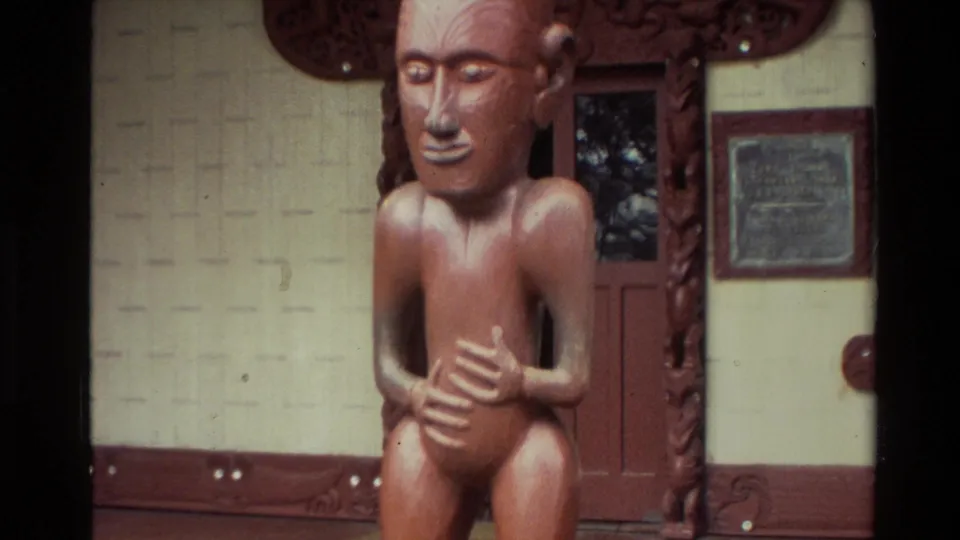 1981 New Zealand home-movie frame of a carved seated figure near a decorated doorway, wall panels, and a plaque-like sign.