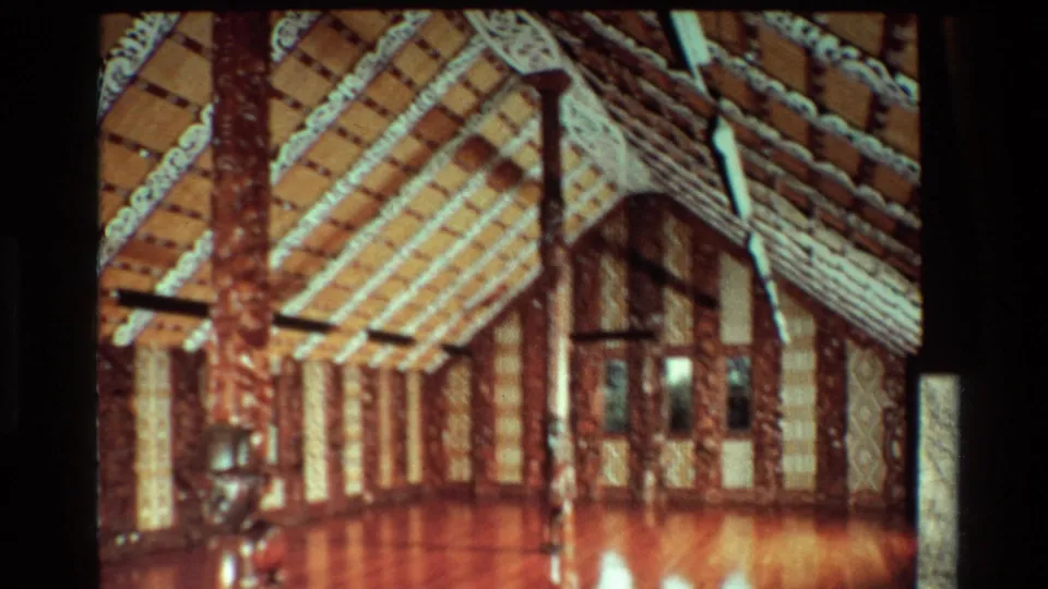 1981 New Zealand home-movie view of a Māori meeting house interior with carved supports, decorated rafters, and warm floor reflections.