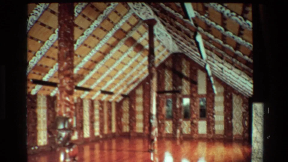 1981 New Zealand 8mm still of a Māori meeting house interior, with carved posts rising toward a patterned ceiling.