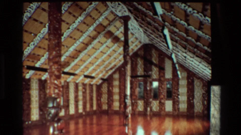 1981 New Zealand frame showing the carved and patterned interior of a Māori meeting house, seen from a handheld visitor perspective.