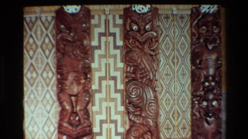 1981 New Zealand home-movie frame showing Maori carved figures and woven panels with soft focus and dark film edging.