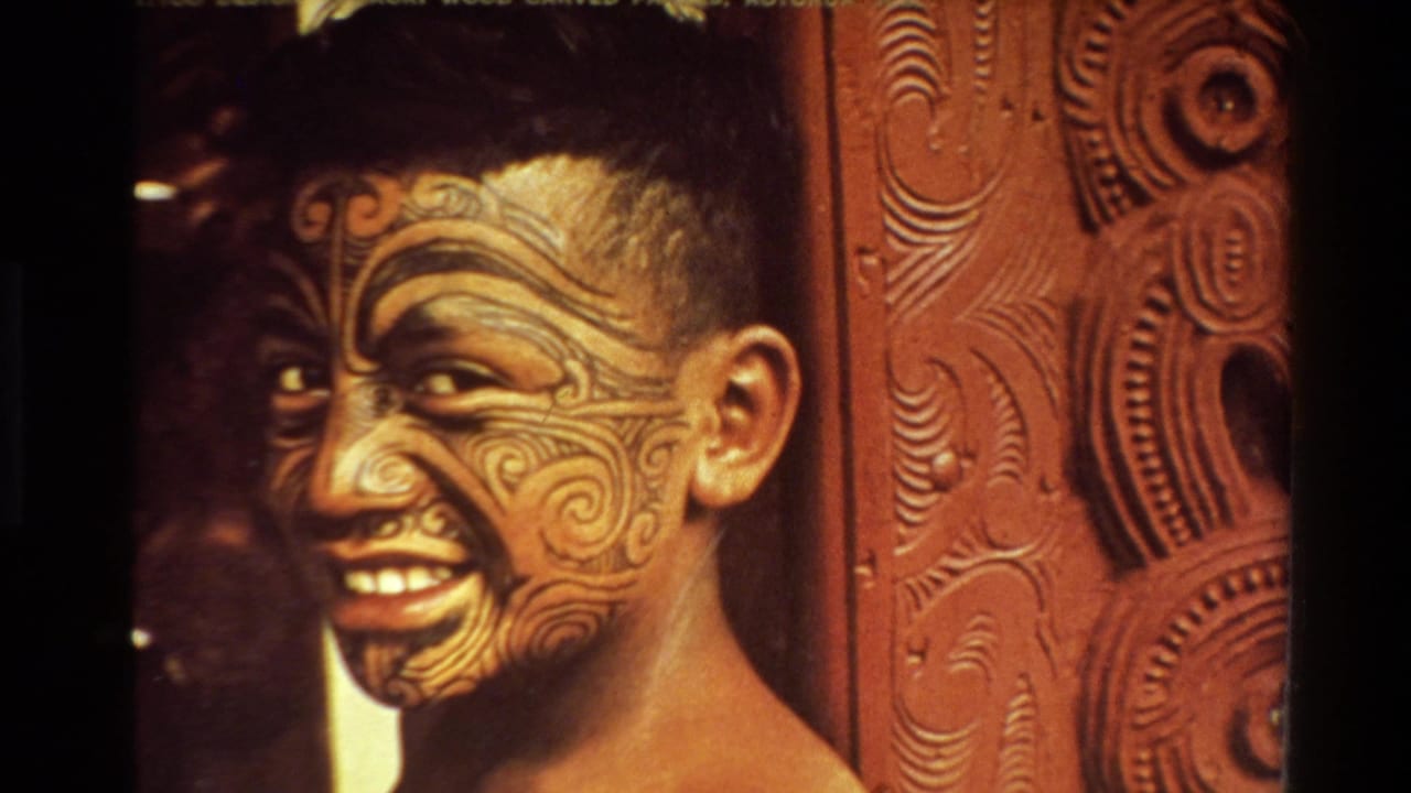 Young person smiling with facial moko-style designs beside carved wooden panels.
