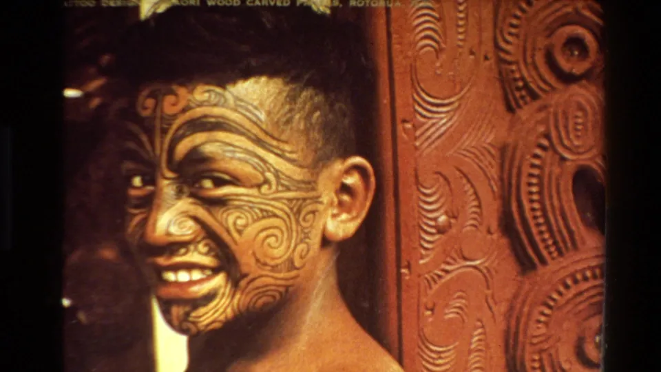 A close 8mm frame shows a smiling young person with moko-style facial designs beside ornate carved wood in New Zealand footage.