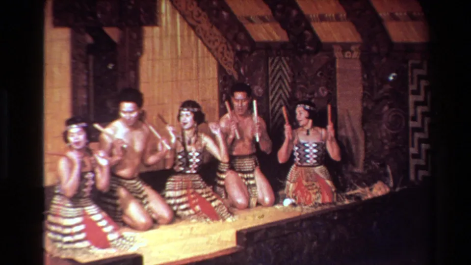 Performers in patterned attire kneel with rhythm sticks inside a carved interior, as seen in 1981 New Zealand home-movie footage.