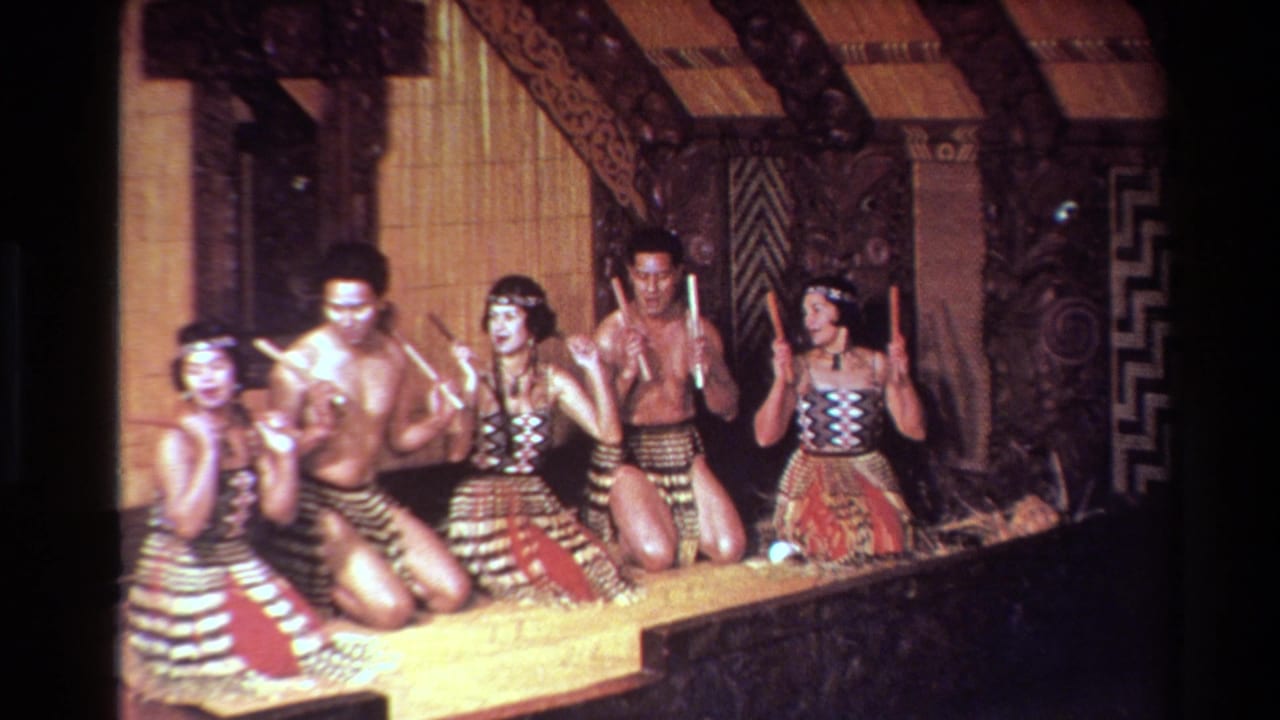 1981 New Zealand 8mm frame of Maori cultural performers kneeling on a carved wooden stage with patterned tops, woven skirts, and rhythm sticks.