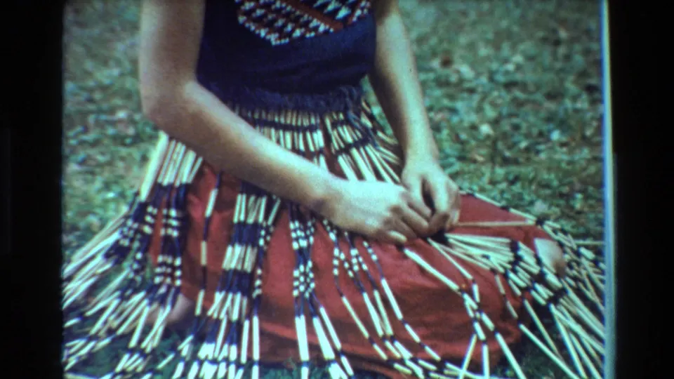 Close 1981 New Zealand 8mm view of a woman’s hands working long light-colored strands on a red and black patterned skirt outdoors.