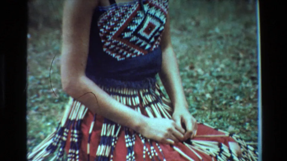 1981 New Zealand 8mm close-up of a person in a patterned top arranging fiber or reed strands over traditional dress on grass.