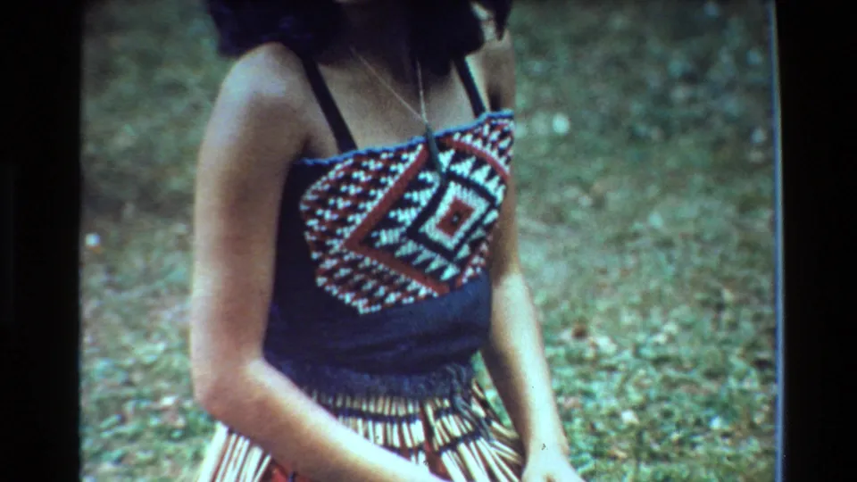 1981 New Zealand 8mm frame showing a woman from shoulders to waist in a patterned top, necklace, and woven skirt against a grassy background.