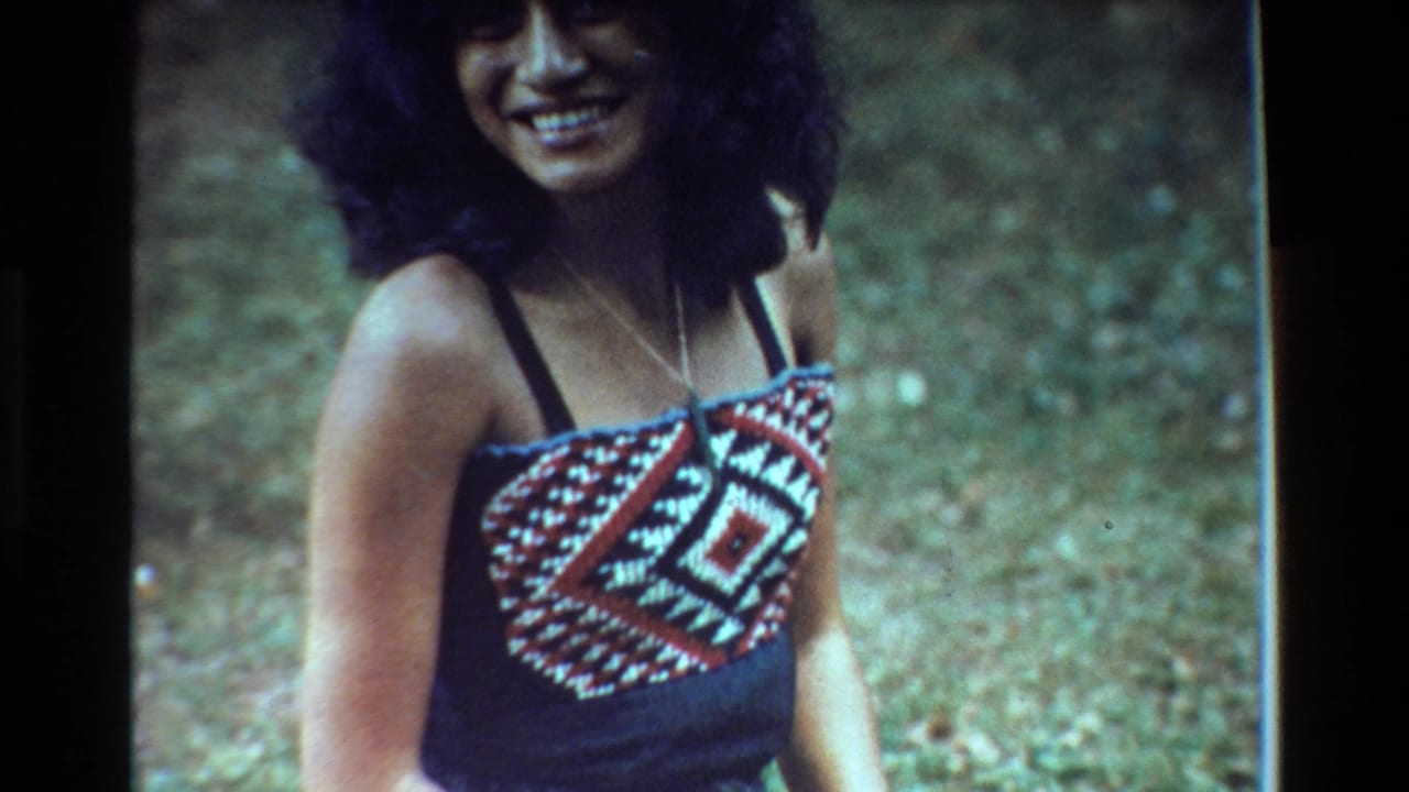 Young woman with dark curly hair smiles outdoors in a patterned sleeveless top.