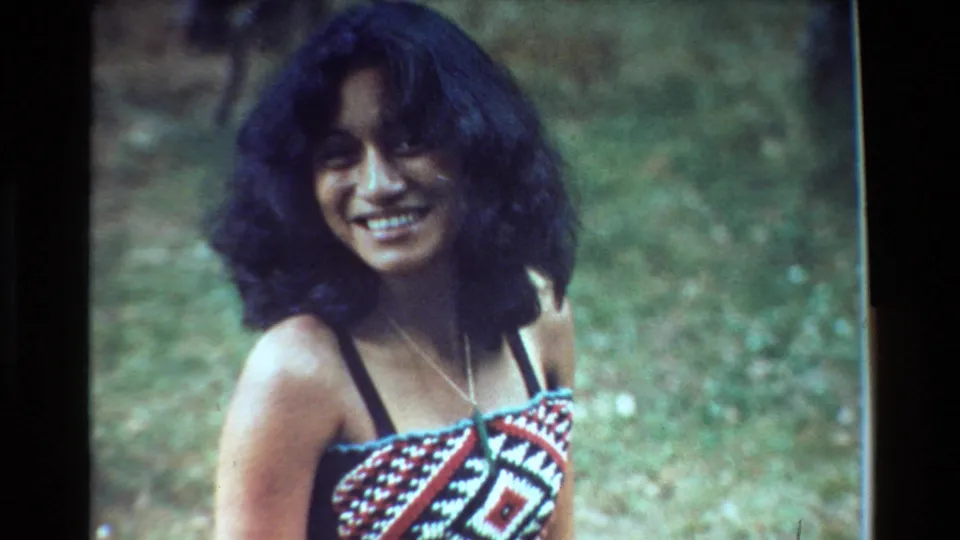 The woman smiles directly toward the camera in an outdoor New Zealand 1981 home-movie shot, with grass behind her and a necklace visible.