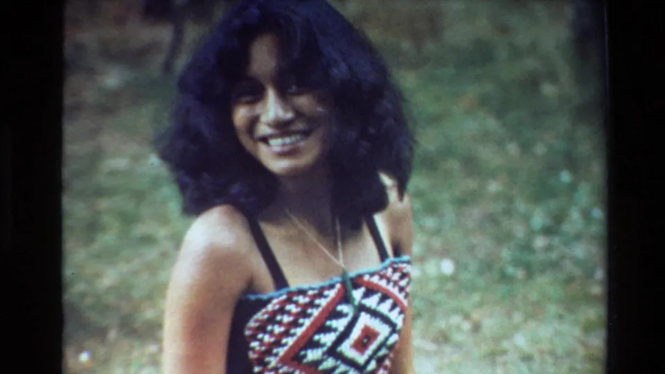 A close home-movie frame shows the smiling woman seated or kneeling on grass, wearing a patterned red, white, and black top in New Zealand in 1981.