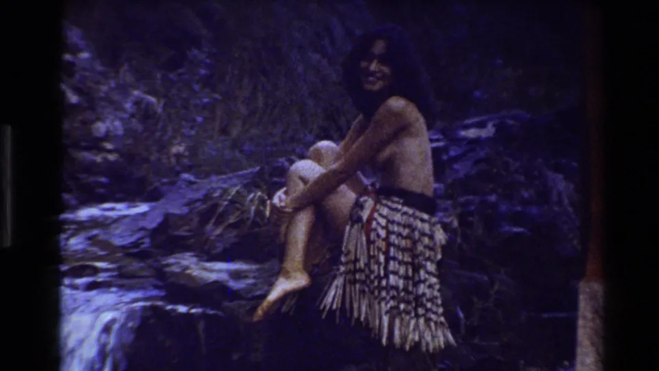 The woman sits on rocks near flowing water in New Zealand, wearing a woven skirt and smiling toward the camera.