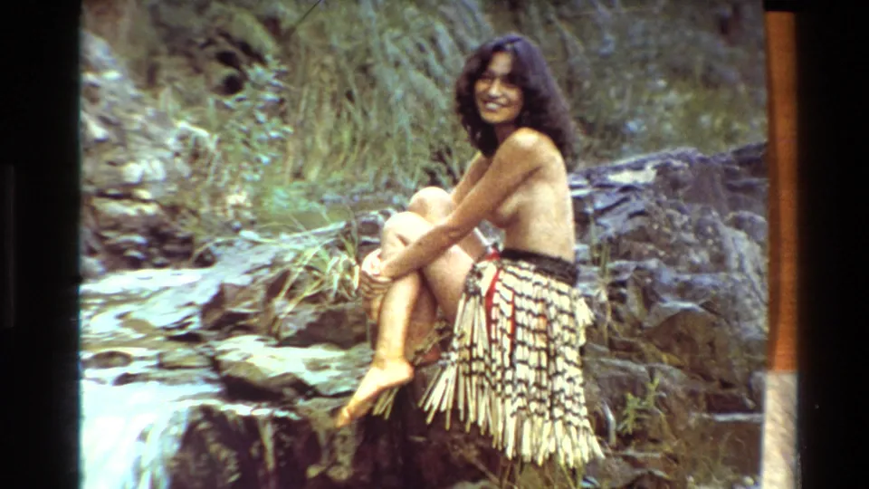 A 1981 New Zealand frame shows the woman seated on rocks by a small waterfall, smiling in a woven skirt.