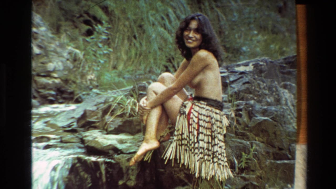 In 1981 New Zealand footage, a smiling topless woman in a feathered skirt sits on rocks near a forest stream.