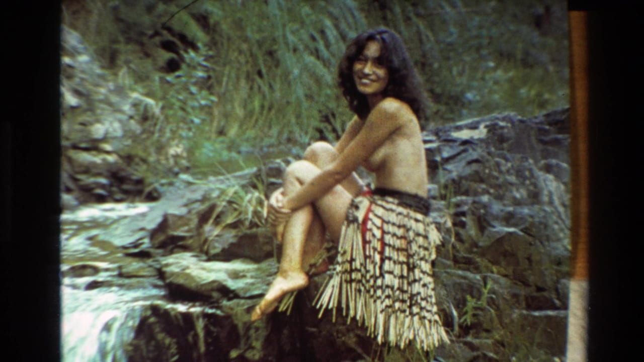 Smiling woman in Maori-style dress sits informally beside a forest stream in 1981 New Zealand home-movie footage.