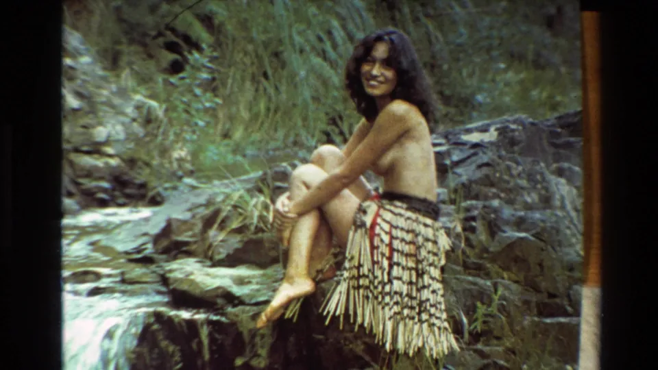 Smiling woman in Maori-style dress sits informally beside a forest stream in 1981 New Zealand home-movie footage.