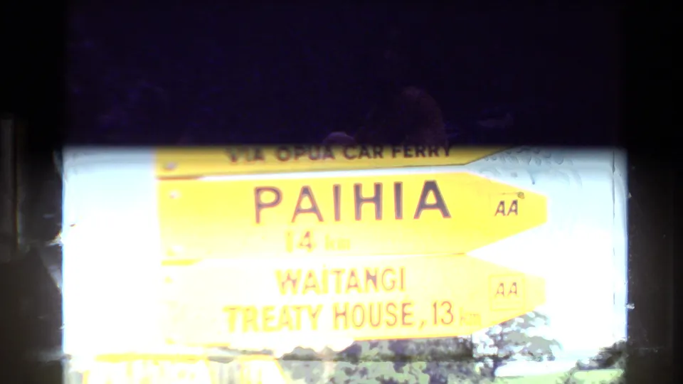 Partly overexposed road sign in New Zealand points toward Paihia and Waitangi Treaty House, with AA marks visible.