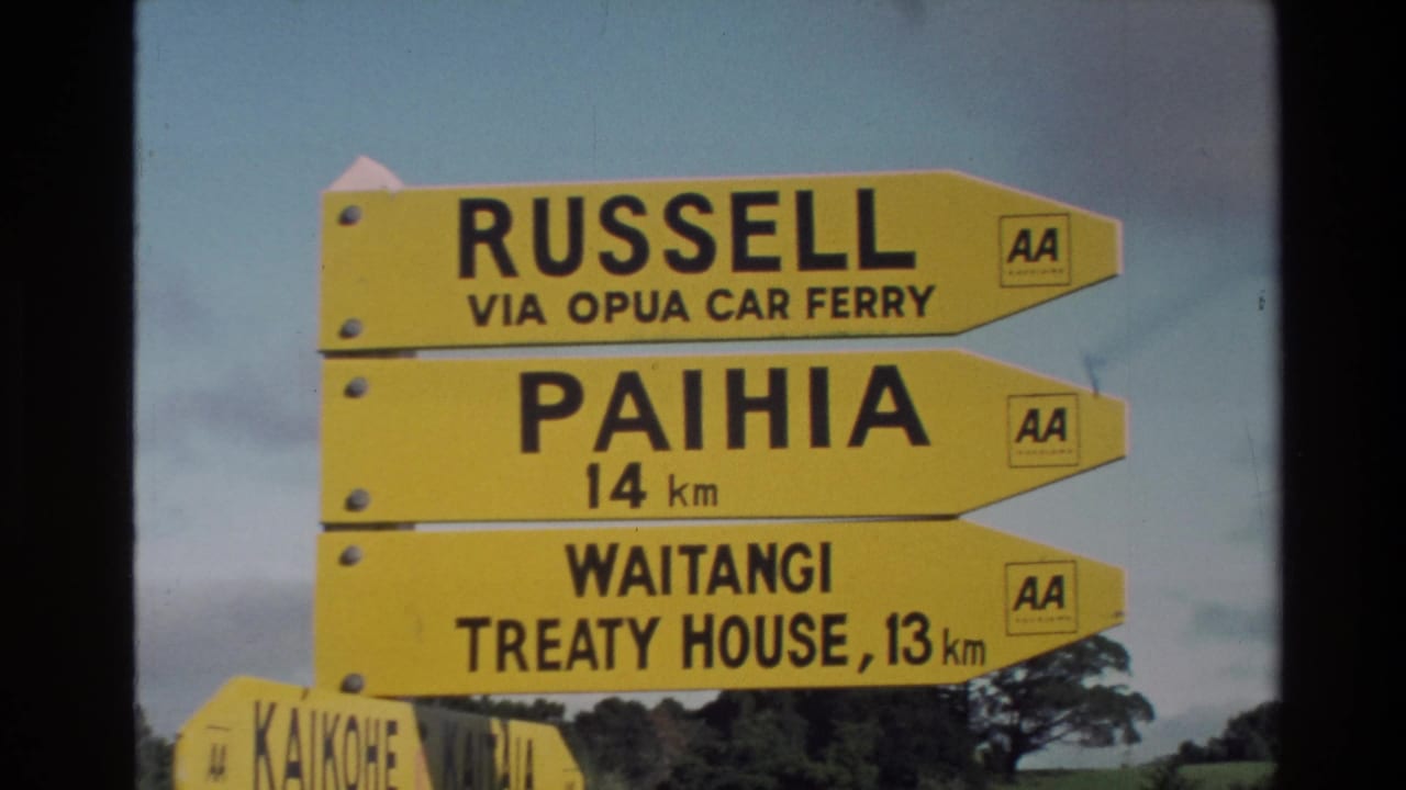 1981 New Zealand home-movie frame showing yellow AA road signs for Russell, Paihia, and Waitangi Treaty House under a cloudy sky.
