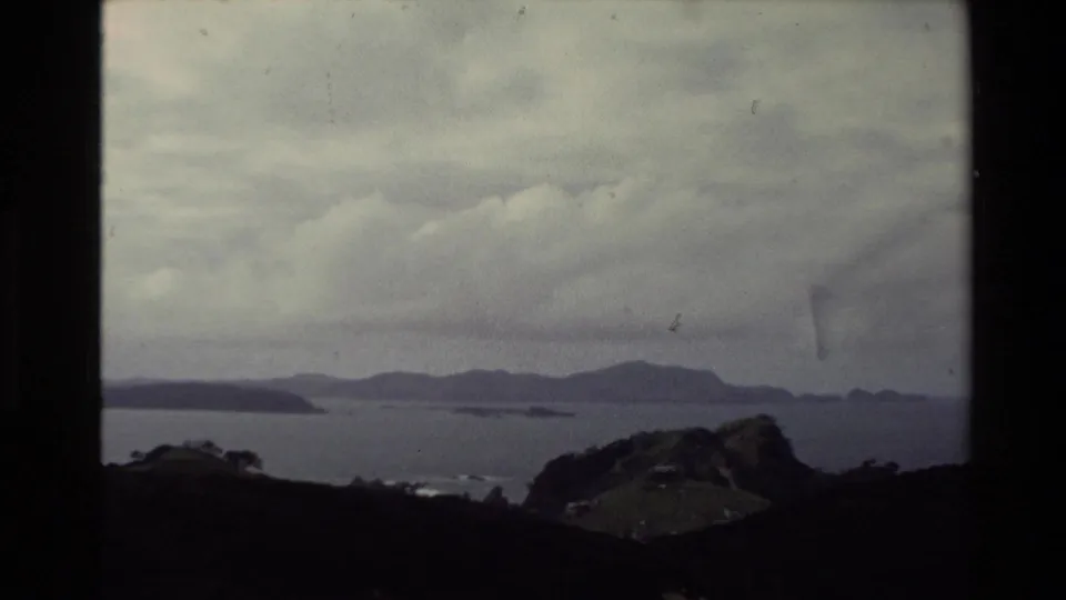 1981 New Zealand home-movie frame showing a misty coastal or bay view with dark foreground hills and distant landforms beneath heavy clouds.