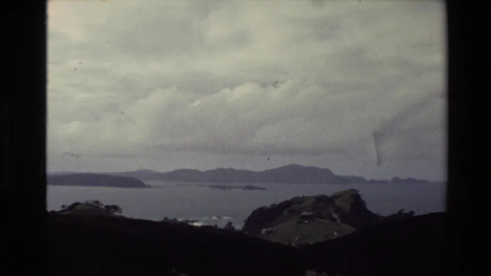 New Zealand 1981 travel frame looking across a calm bay or coast toward distant islands or mountains under a thick overcast sky.