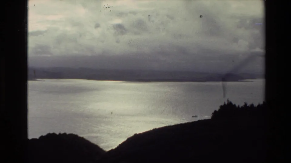 1981 New Zealand 8mm view across a broad body of water, with distant shoreforms and dark hill slopes in the foreground.