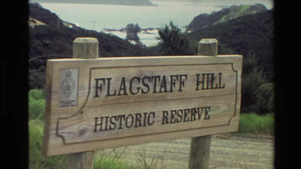1981 New Zealand frame showing the wooden Flagstaff Hill Historic Reserve sign, with coastal vegetation and water beyond.