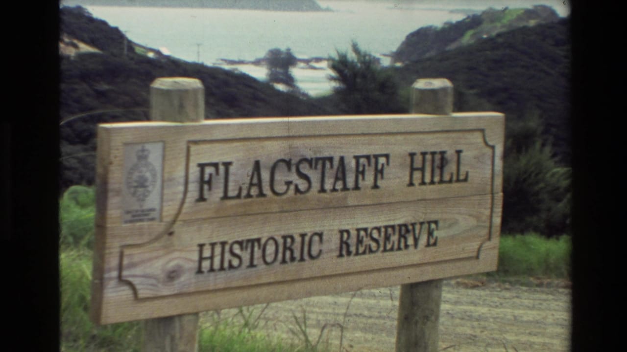 1981 New Zealand home-movie frame showing a wooden Flagstaff Hill Historic Reserve sign with coastline and vegetation behind it.