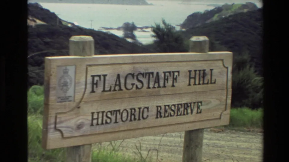 1981 New Zealand home-movie frame showing a wooden Flagstaff Hill Historic Reserve sign with coastline and vegetation behind it.