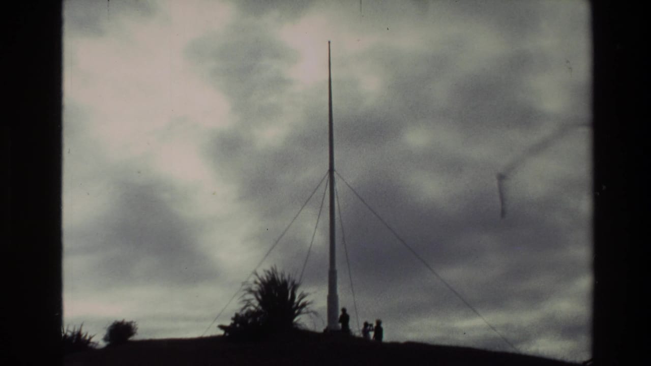 Silhouetted flagstaff with guy wires on a hill under cloudy sky.
